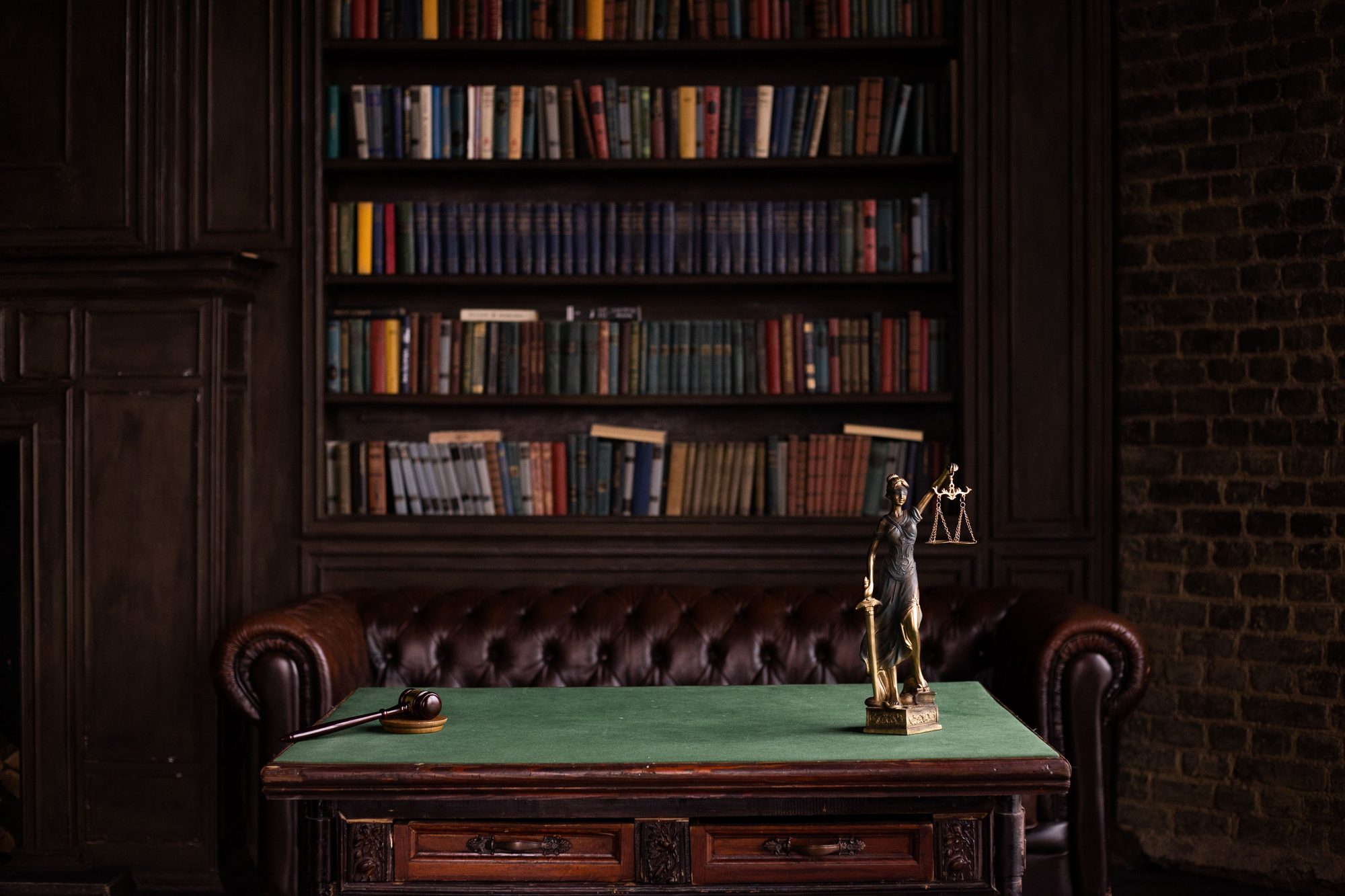 Traditional law office library with a leather chesterfield sofa, shelves of legal volumes, a gavel, and a bronze statue of Lady Justice on an antique desk.
