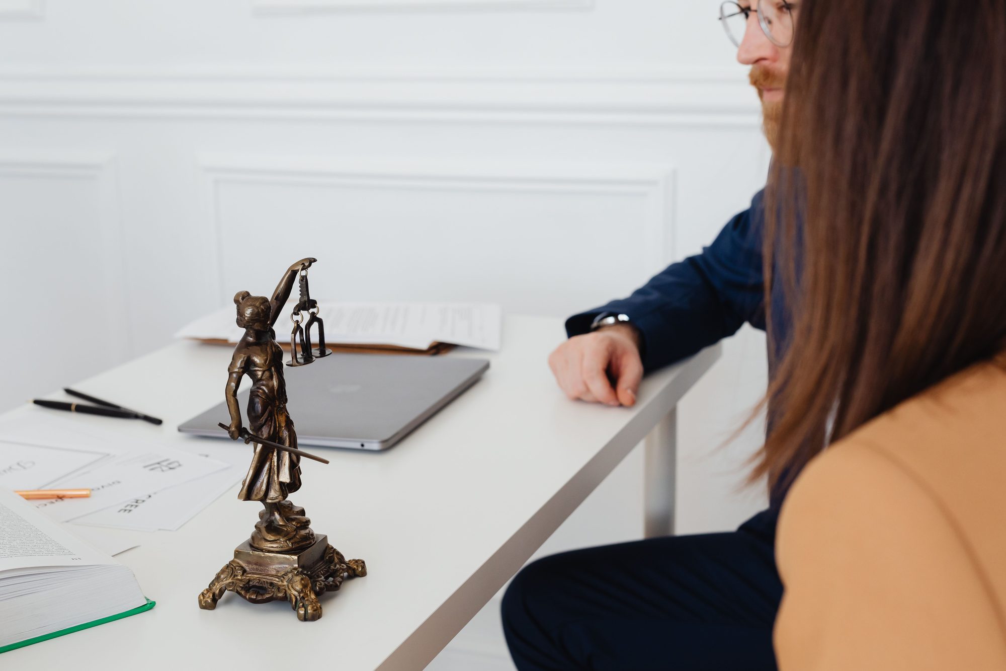 A bronze statue of Lady Justice holding scales on a desk, with two colleagues in business attire reviewing documents in the background.