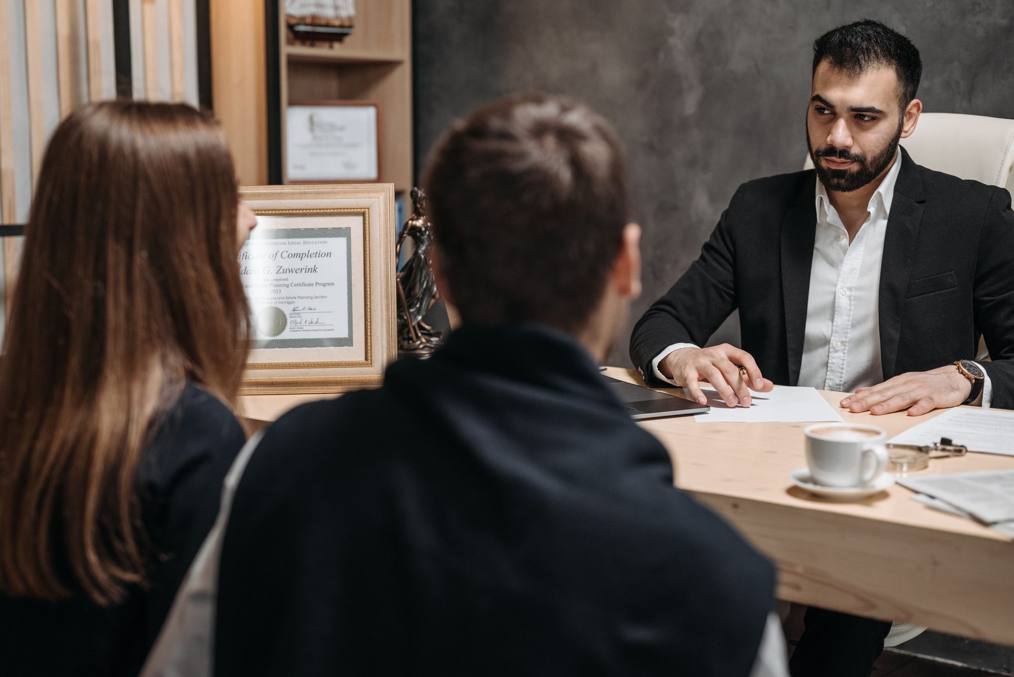 An attorney in a dark suit consulting with two clients at his desk, reviewing documents together in a modern office.
