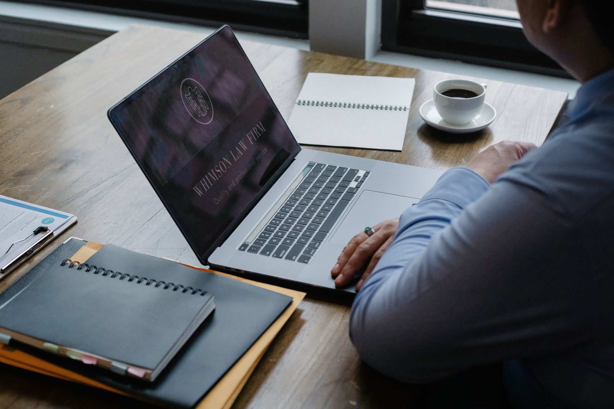 Close-up of a laptop keyboard, a cup of coffee, and an open notebook on a wooden desk, with an attorney's hand resting on the trackpad while preparing a case.